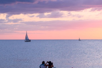 Nokomis Beach: Couple Watches a Sailboat Cruise Against a Pale Pink Sky